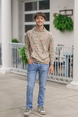A full-length front view of a smiling young man standing on an outdoor concrete patio wearing the Retro Woodlands Performance Hoodie. The lightweight activewear pullover features a vintage-inspired camouflage pattern in warm, earthy shades of tan, sage green, and soft brown. It is styled with a sleek crossover hood, a spacious front kangaroo pocket, light-wash slim-fit denim jeans, and olive-green and white sneakers.