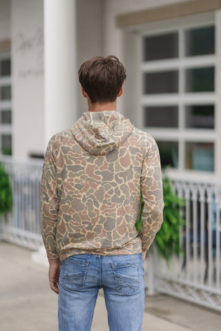 Back view from the waist up of a young man wearing the Retro Woodlands Performance Hoodie, emphasizing the fluid fit and continuous print from behind. The retro-inspired, earth-toned camouflage pattern runs seamlessly across the back panel, hood, and raglan shoulder seams, showing off the relaxed, active-mobility design paired with light-wash denim jeans.