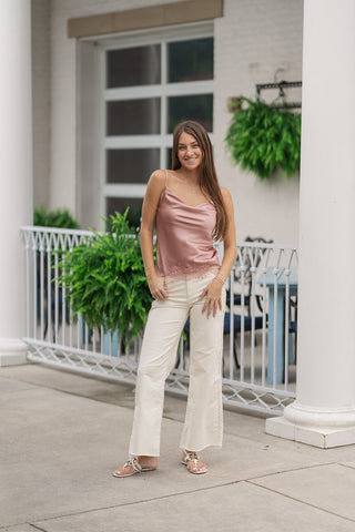 A full-length view of a smiling woman posing on an outdoor patio wearing the Pink Champagne Satin Tank. The sleeveless camisole top features a luxurious satin fabric in a soft pink champagne color with a fluid, draped cowl neckline. It is styled for a casual yet elegant summer look with high-waisted cream-colored wide-leg trousers, flat white leather slide sandals, and a delicate gold cross necklace.