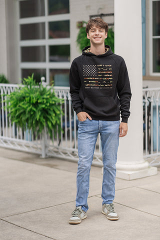 A full-length front view of a smiling young man standing outdoors on a concrete patio wearing the Onyx Camo Flag Hoodie. The long-sleeve black hoodie features a prominent American flag graphic on the chest with camo-filled stripes and a field of white stars, with "SIMPLY SOUTHERN OUTFITTERS" printed below it. The patriotic, rugged look is styled with light-wash slim-fit denim jeans and olive-green and white sneakers in front of a white columned entryway.