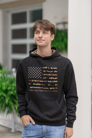 An upper-body portrait view of the Onyx Camo Flag Hoodie highlighting the intricate chest graphic. The solid black pullover design features a built-in hood lined with matching woodland camouflage print fabric. The chest graphic showcases detailed camouflage texturing within the flag's horizontal stripes, sitting above a front kangaroo pocket where a small embroidered dog silhouette logo is subtly visible.