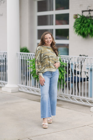 A full-length front view of a smiling woman standing on an outdoor patio wearing the Dear John Riviera Camo Trenton pullover. This lightweight cotton-blend knit top features an artistic camo print in soft sage, olive, and khaki tones. The relaxed, slouchy silhouette is styled with light-wash wide-leg jeans and cream platform wedge sandals in front of a white columned building.