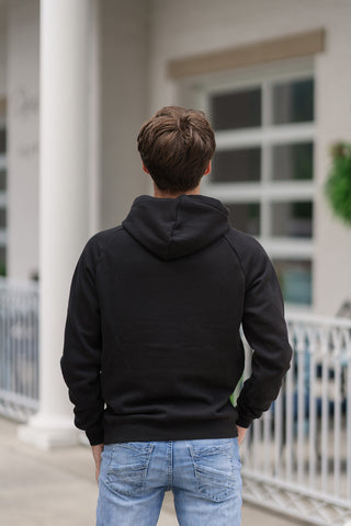 Back view from the waist up of a young man wearing the Onyx Camo Flag Hoodie, emphasizing the clean, minimalist look from behind. The solid black base fabric offers a structured yet comfortable fit with raglan-style shoulder seams, showing the attached hood resting neatly at the upper back. The hoodie is paired with light-wash blue jeans on an outdoor patio.