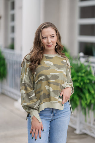 An upper-body portrait view of the Dear John Riviera Camo Trenton pullover highlighting its cozy, lightweight texture. The design features a flattering, wide scoop neckline and comfortable drop-shoulder sleeves. The model poses with one hand near her waist, showcasing the relaxed, non-clinging drape of the earthy camo pattern.