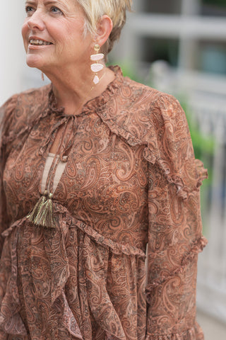 A portrait-style close-up shot focusing on the intricate bodice details of the Boho Breeze Chiffon Mini dress. The lightweight printed chiffon fabric is gathered at the elasticized waist, displaying the plunging V-neckline framed by delicate ruffle trims and a keyed tie closure with shimmering gold-threaded tassels. The woman looks upward and smiles, highlighting her gold and light pink stone geometric drop earrings.