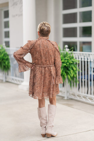Back view of a woman standing on a concrete patio wearing the Boho Breeze Chiffon Mini dress, emphasizing the fluid movement and ruffle layers from behind. The earthy paisley print runs seamlessly down the back of the dress, accented by cascading ruffle overlays along the shoulders, elasticized waistline, and tiered skirt hem. The woman poses with one hand on her hip, showcasing the sheer long sleeves and flared trumpet cuffs, paired with tall cream suede boots.