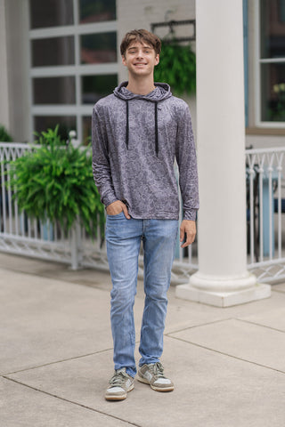 A full-length front view of a smiling young man standing on an outdoor patio wearing the Shadow Camo Performance Hoodie. The lightweight activewear top features a sleek, tonal grey camouflage pattern throughout its smooth performance fabric. It is styled with a crossover-style neckline, a relaxed built-in hood, and contrasting black drawstrings. The casual athletic ensemble is paired with light-wash slim-fit denim jeans and grey-green accented sneakers in front of a white columned background.