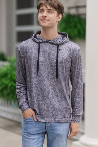An upper-body portrait view of the Shadow Camo Performance Hoodie showcasing its high-performance details. The smooth, multi-tone grey allover camouflage print covers the body and long sleeves, which are finished with clean, self-fabric cuffs. A small, subtle black dog silhouette logo is embroidered on the left chest. The man poses with one hand casually tucked into his front jean pocket.