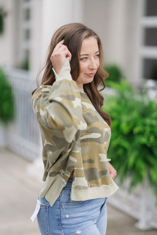 A close-up side profile view of the Dear John Riviera Camo Trenton pullover focusing on its dimensional design details. This angle highlights the thick ribbed trim along the high-low hemline and the subtle side-slit accents that allow the fabric to drape beautifully. The model raises her hand, putting the long sleeves with their unique split-cuff wrist slit details on display.