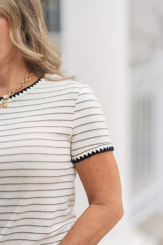A detailed close-up shot focusing on the artisanal shoulder and sleeve elements of the Scalloped Edge Sweetheart Tee. The image highlights the clean horizontal alignment of the thin black micro-stripes on the soft cream fabric, along with the intricate, textured black crocheting stitch that trims the collar and the edge of the short sleeve cuff.