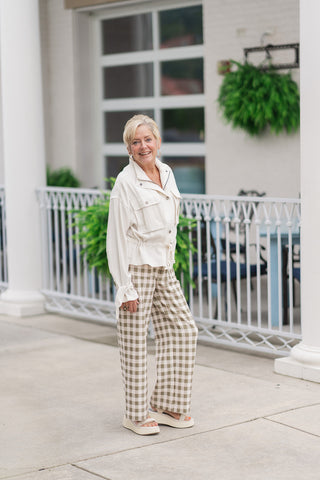 A woman smiling and posing on an outdoor patio wearing the Canyon Clay Cargo Jacket, a lightweight utility jacket in a neutral ecru tone. The design features a structured stand collar, pleated cargo chest pockets with snap closures, and a gathered drawstring waist tied at the front. The look is styled for a casual boutique aesthetic with olive-green and cream gingham check wide-leg pants and cream-colored platform slide sandals.