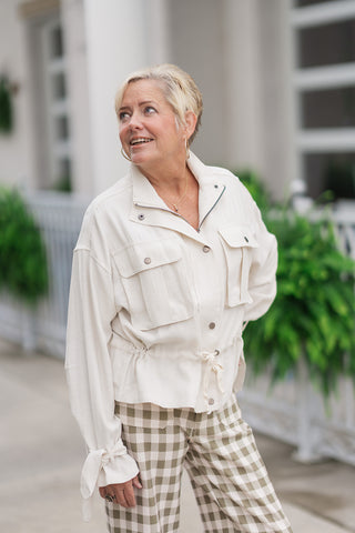 A mid-shot view of the Canyon Clay Cargo Jacket highlighting its utility-inspired structure and relaxed drape. The ecru-colored woven fabric features drop-shoulder seams, a partial-zip front opening, and unique long sleeves finished with self-tie ribbon cuffs knotted at the wrists. The adjustable drawstring waist is subtly cinched to define her natural shape over matching checkered trousers.