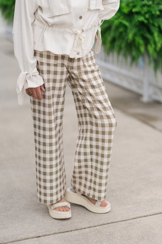 A close-up mid-shot focusing on the design details of the Olive Grove Wide-Leg Pants from the waist down. The image highlights the soft drape of the olive-green and cream gingham print knit fabric. Structured details are visible, including the secure zip-fly single button front closure and eye-catching front patch pockets at the waist, styled with chunky off-white flatform sandals.