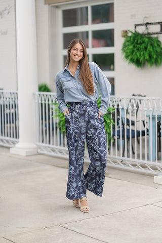 A full-length view of a smiling woman posing on an outdoor patio in the Metropolitan Chambray Top. The lightweight washed tencel shirt is featured in a classic light blue denim look and is styled cleanly tucked into high-waisted navy blue wide-leg trousers adorned with a light blue floral print. The relaxed outfit is finished with beige flat woven slide sandals and a delicate gold cross necklace.