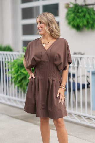 A portrait-style mid-shot of the Espresso Essence Pocket Dress highlighting its contemporary, tailored elements. The lightweight espresso brown knit fabrication shapes the casual drape of the short dolman sleeves and clean V-neckline. The structural vertical pleating along the waist panel creates a defined silhouette, accessorized beautifully with a gold coin charm necklace and stacked metallic bracelets.
