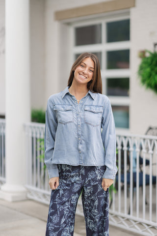 An upper-body portrait view of a smiling woman wearing the Metropolitan Chambray Top loose and untucked to highlight its fluid, boxy drape. The soft tencel fabric displays a beautiful light blue denim wash with subtle vintage-inspired fading along the seams, a structured pointed collar, a button-down placket, and dual chest flap pockets.