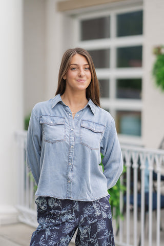 A detailed close-up shot focusing on the front bodice elements of the Metropolitan Chambray Top. The image highlights the smooth, silky-soft texture of the washed tencel material, the classic pointed collar, and two functional chest flap pockets with central button closures. The shirt features matching light-toned button details along the front placket, styled over dark floral wide-leg trousers.
