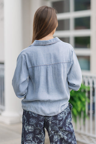 Back view from the waist up of a woman wearing the Metropolitan Chambray Top, showcasing the clean design of the back panel. The light blue tencel material drapes beautifully, emphasizing a structured horizontal back yoke seam and a classic center box pleat that adds fluid movement to the relaxed, long-sleeve silhouette.