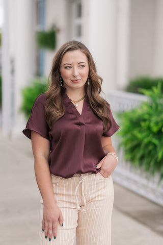 A portrait-style mid-shot highlighting the front design details of the Plum Velvet Dreams Top. The premium crinkle satin fabric drapes elegantly, showcasing the modern V-neckline, minimalist mandarin-style partial collar, and a subtle centered front pleat. The woman poses with one hand in her pocket, accessorized with a gold link necklace with a baroque pearl pendant and stacked bracelets.