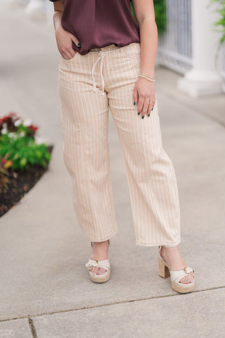 A mid-shot view from the waist down focusing on the front construction of the Toasted Almond Barrel Pants. The cream and taupe pinstriped fabric highlights distinct vertical center seams running down the front of each leg and a high-rise waistline. The trousers are styled with a removable, adjustable matching cream rope tie belt knotted at the center and paired with gold-buckled platform sandals.