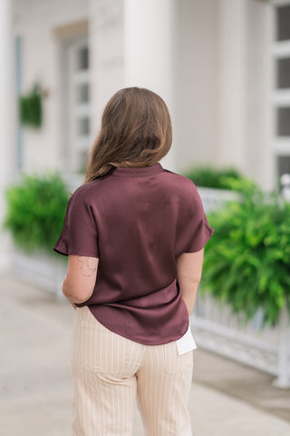 Back view from the waist up of a woman wearing the Plum Velvet Dreams Top, emphasizing its relaxed drop-shoulder silhouette and short cap sleeves. The rich plum satin fabric catches the light, showcasing its texture and the fluid flow of the curved high-low hemline as it rests untucked against the waistband of her pinstriped trousers.