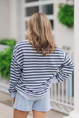 Back view from the waist up of a woman wearing the Z Supply Everyday Striped Crew, emphasizing the continuous drape of the horizontal stripe pattern across the reverse side. The slouchy drop-shoulder sleeves and relaxed back panel highlight the clean, matching alignment of the navy and light blue stripes, finished with a thick ribbed knit hem at the waistband of her light blue shorts.