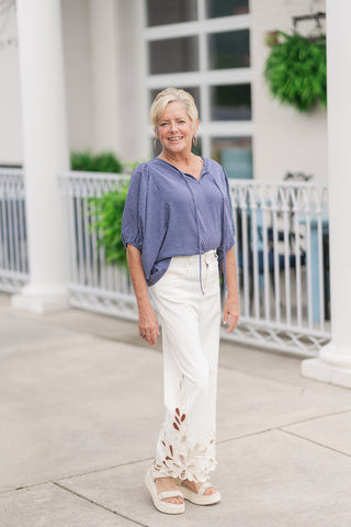 A full-length front view of a smiling woman standing on an outdoor patio wearing the Free Spirit Sailor Blouse. The lightweight, short-sleeve top features a timeless vertical pinstripe pattern in alternating shades of navy blue and soft cream, styled with a modern split V-neckline and relaxed dolman sleeves. The blouse is styled for an elevated, breezy coastal look with a front tuck into high-waisted white wide-leg trousers featuring intricate beige floral eyelet cutouts along the bottom hem.