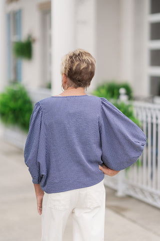 Back view from the waist up of a woman wearing the Free Spirit Sailor Blouse, emphasizing the clean, continuous flow of the vertical pinstripe pattern across the reverse side. This angle showcases the generous drape of the relaxed dolman sleeves and the structured line of the boat-style rear collar, paired with high-waisted white pants on an outdoor concrete patio.