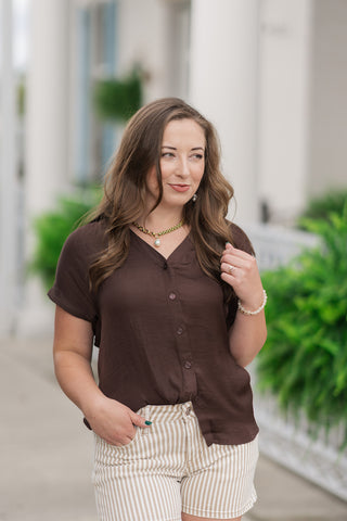 An upper-body portrait view of the Hazelnut Horizon Top highlighting the texturized satin-blend fabric. The design features a classic pointed shirt collar framing a flattering open V-neckline with a tonal button placket down the front. The short cuffed cap sleeves show a subtle rolled-edge finish, accessorized with a gold link necklace with a baroque pearl drop and neutral beaded bracelets.