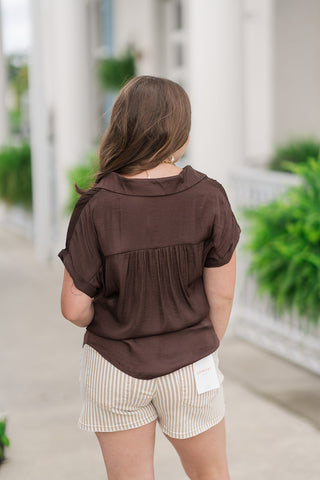 Back view from the waist up of a woman wearing the Hazelnut Horizon Top, showcasing the fluid drape of the relaxed silhouette. The chocolate brown satin-feel fabric features a gathered back yoke that adds rich dimension and movement across the shoulders. The blouse is left untucked to display the straight hemline against the waistband of her striped neutral shorts.