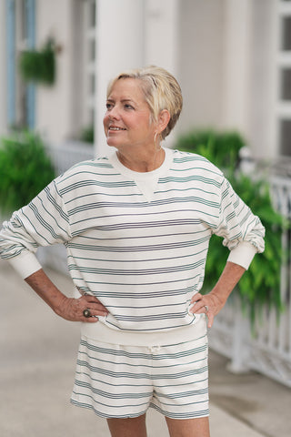 An upper-body portrait view of the Z Supply Stand Out Sweatshirt showcasing its relaxed, slouchy fit. The design features dropped shoulders, comfortable long sleeves, and a classic crew neckline accented with a vintage-inspired sporty triangle stitch detail at the center collar. The woman poses with her hands on her hips, looking off to the side, styled with matching cream and black striped loungewear shorts.