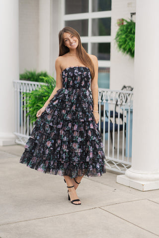 A full-length front view of a smiling woman standing on an outdoor patio wearing the Cascading Garden Strapless Dress. The elegant, ankle-length maxi dress features a sheer black tulle overlay adorned with a moody, vibrant floral print in shades of purple, pink, and green. The dress showcases a strapless silhouette with structured, tiered ruffling that cascades down the skirt for soft volume and fluid motion. It is styled for a romantic summer garden party look with black strappy block-heel sandals and a de