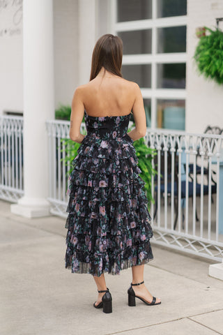 Back view of the Cascading Garden Strapless Dress, showcasing the dress's tiered ruffle layout and fit from behind. The strapless bodice features a secure, comfortable smocked back panel in the matching dark floral print. The sheer, layered ruffles of the tulle skirt cascade down to a graceful maxi length, styled with black ankle-strap block-heeled sandals on a concrete patio.