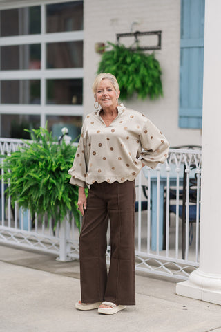 A full-length front view of a smiling woman standing on an outdoor patio wearing the Sweet Wave Collared Blouse. The lightweight woven blouse showcases a shimmering sage-champagne hue adorned with a textured bronze and brown polka dot print. It features an elegant standing collar, a V-neck placket, dramatic 3/4 puff sleeves, and a beautifully detailed scalloped hemline. The top is styled for a sophisticated daytime look with dark brown wide-leg trousers and cream platform slide sandals.