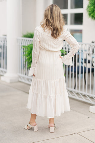 Back view of the Victorian Romance Midi Dress showcasing the clean, elongated silhouette and beautiful movement of the tiered midi skirt. This view emphasizes the gathered, elasticized waistline that defines the shape comfortably, the subtle vertical textured fabric, and the flowing layers of the skirt. The woman is posed looking away, showing the back of the ruffled neckline and long sleeves.