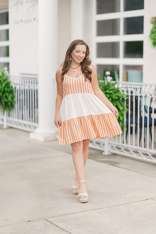 A full-length view of a woman smiling and walking forward in the Sunkissed Sherbet Tiered Dress, a playful color-blocked babydoll mini dress. The design showcases textured vertical stripes in a warm sherbet orange and cream hue on the top and bottom tiers, contrasted by a solid cream middle tier. She is holding the lightweight skirt slightly to the side, showcasing its breezy, tiered silhouette. The outfit is styled for a chic summer look with cream espadrille wedge sandals, a gold chain pearl necklace,.
