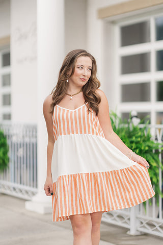 A portrait-style mid-shot of the Sunkissed Sherbet Tiered Dress, highlighting the flattering V-neckline with a clean, banded border and slender, adjustable spaghetti straps. The textured woven fabric features a warm orange-and-cream striped pattern on the bodice, transitioning into a solid cream tiered panel at the waist. The woman is looking to the side, accessorized with a chunky gold chain necklace featuring a baroque pearl drop and matching pearl earrings.