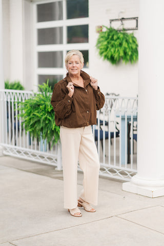 A full-length front view of a smiling woman standing on an outdoor patio wearing the Mocha Muse Bomber Jacket. The jacket is rendered in a deep, rich mocha brown earth tone and features a high stand collar, military-inspired shoulder tab details, a button-down front, and a gathered hem that creates a distinct blouson effect. The utility-inspired look is styled with tailored cream wide-leg trousers, beige open-toe slide sandals, and gold hoop earrings.
