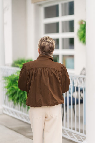 Back view from the waist up of a woman wearing the Mocha Muse Bomber Jacket, highlighting the gathers along the back yoke that provide fluid movement and structural dimension. The rich mocha brown utility fabric drapes cleanly down to a gathered elastic hem, creating a soft blouson-style shape across the lower back. The jacket is paired with high-waisted cream trousers on an outdoor patio setting.