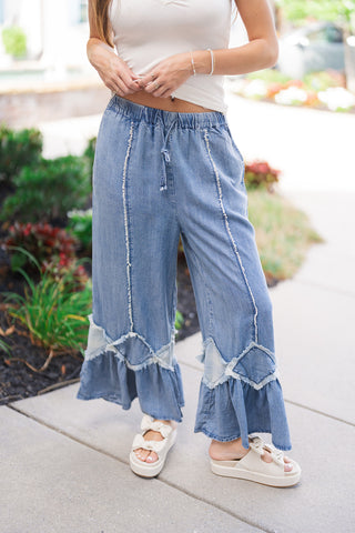 A detailed close-up shot focusing on the front construction of the Flirty Flare Ruffle Denim Pants. The view highlights the gathered elastic drawstring waist, prominent vertical raw-edge frayed seams running down each leg, and the geometric diamond patch design that transitions into wide, ruffled flounce hems near the ankles.