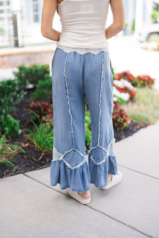 A rear view of the Flirty Flare Ruffle Denim Pants showing the back fit and design details. The shot highlights the elasticized waistband, vertical raw-edge seams extending down the back of the legs, and the low-set diamond patchwork piecing leading into the dramatic, airy ruffle-trimmed hems.