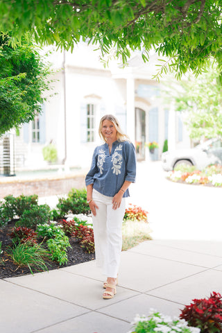 A smiling blonde woman posing outdoors in the Blossom & Blue Denim Tunic—a medium-wash blue denim tunic top featuring large white floral felt appliqué artwork across the front chest, a split notch neckline with white piping, and 3/4-length sleeves with subtle shoulder gathers. She has styled the top for a fresh daytime outfit paired with crisp white high-waisted trousers and neutral platform sandals.