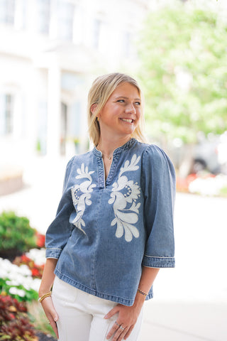 A mid-length portrait view highlighting the front details of the Blossom & Blue Denim Tunic. The shot emphasizes the bold contrast of the large-scale white floral appliqué framing the split v-neckline, the subtle contrast piping along the band collar and sleeve cuffs, and the soft, relaxed drape of the lightweight denim top tucked loosely against white pants.