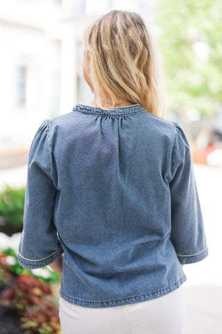 Back view of the Blossom & Blue Denim Tunic showcasing the gentle gathered detail along the back center collar and shoulders for subtle volume. The image highlights the clean, relaxed silhouette of the medium-wash blue denim, the piped edge along the collar, and the 3/4 sleeve hem detailing.