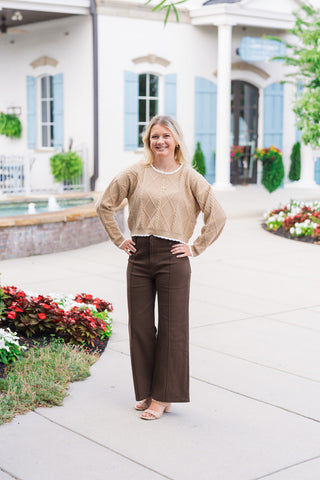 A blonde woman smiling outdoors with her hands on her hips while wearing the Toffee Lattice Scalloped Sweater—a warm toffee-beige cropped cable-knit pullover featuring a raised diamond lattice pattern and delicate white scalloped trim along the crew neckline, wrist cuffs, and hem. Styled for a cozy autumn look paired with high-waisted dark brown wide-leg trousers and woven neutral block heels.