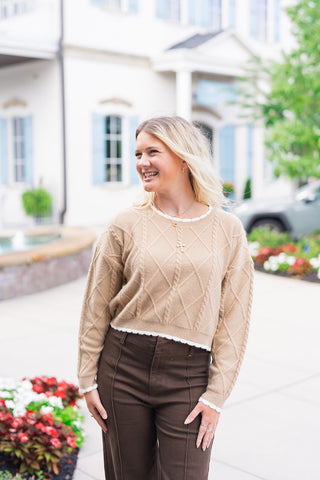 A portrait view of the Toffee Lattice Scalloped Sweater highlighting the raised diamond cable-knit texture and sweet, crisp white scalloped trim. The detail shows the crew neck fit, dropped shoulder seams, and the cropped ribbed hem meeting the waistband of dark brown pants, complemented by layered gold pendant necklaces.