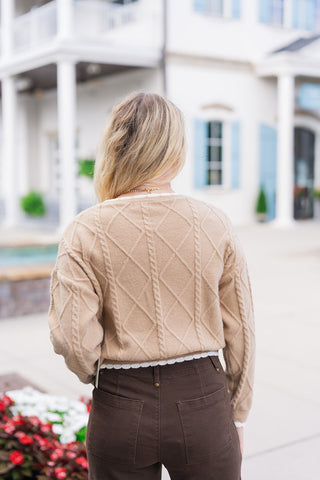 Back view of the Toffee Lattice Scalloped Sweater displaying the continuous diamond cable-knit lattice design running across the shoulders, back, and sleeves. The shot emphasizes the gentle cropped length, white scalloped hem detail, and relaxed shoulder fit paired with high-waisted dark brown pants featuring back patch pockets.