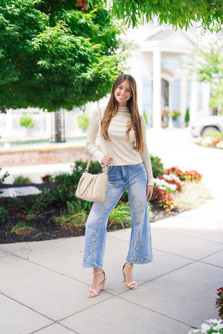 Full-length view of a smiling woman posing outdoors in the Desert Rose Border Jeans—medium-wash, high-waisted denim pants featuring soft fading and intricate ivory floral cross-stitch embroidery along the legs and hem. Styled for a casual daytime look with a cream long-sleeve knit top, a beige shoulder bag, and nude ankle-strap block heel sandals.