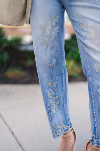 Detailed close-up shot focusing on the lower leg and fabric texture of the Desert Rose Border Jeans. It highlights the fine, cross-stitch style floral embroidery in muted ivory and beige threads contrasting against the sun-washed blue denim, leading down to the decorative hem trim paired with nude ankle-strap heels.