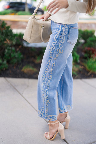 Side profile view of the Desert Rose Border Jeans emphasizing the flattering high-rise waist and back pocket detail. The view showcases the continuous ornate scrollwork embroidery running directly down the outer side seam and wrapping around the bottom cropped hemline.
